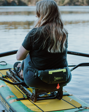 A woman sits on a paddleboard on a lake, viewed from behind. She is wearing a black t-shirt and black pants, with a black waist personal flotation device holding a yellow-green item.