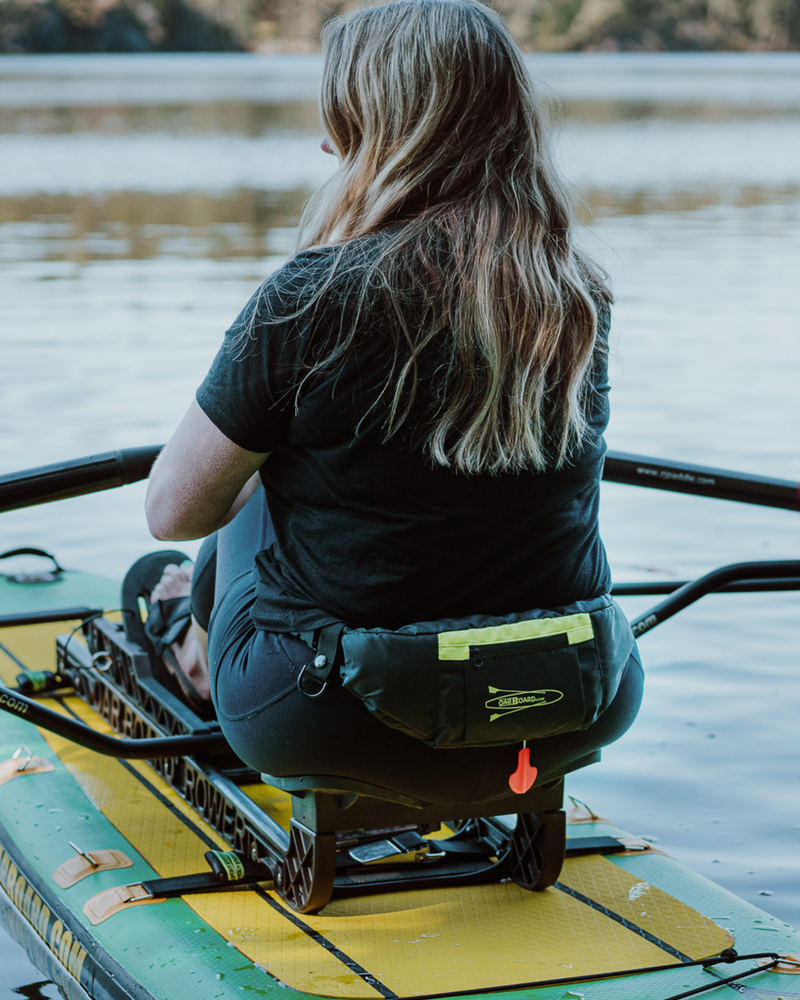 A woman sits on a paddleboard on a lake, viewed from behind. She is wearing a black t-shirt and black pants, with a black waist personal flotation device holding a yellow-green item.