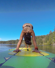 A woman with tattoos on her back and neck is doing a yoga downward dog pose on a green and yellow paddleboard on a calm lake with a forest shoreline under a clear blue sky.