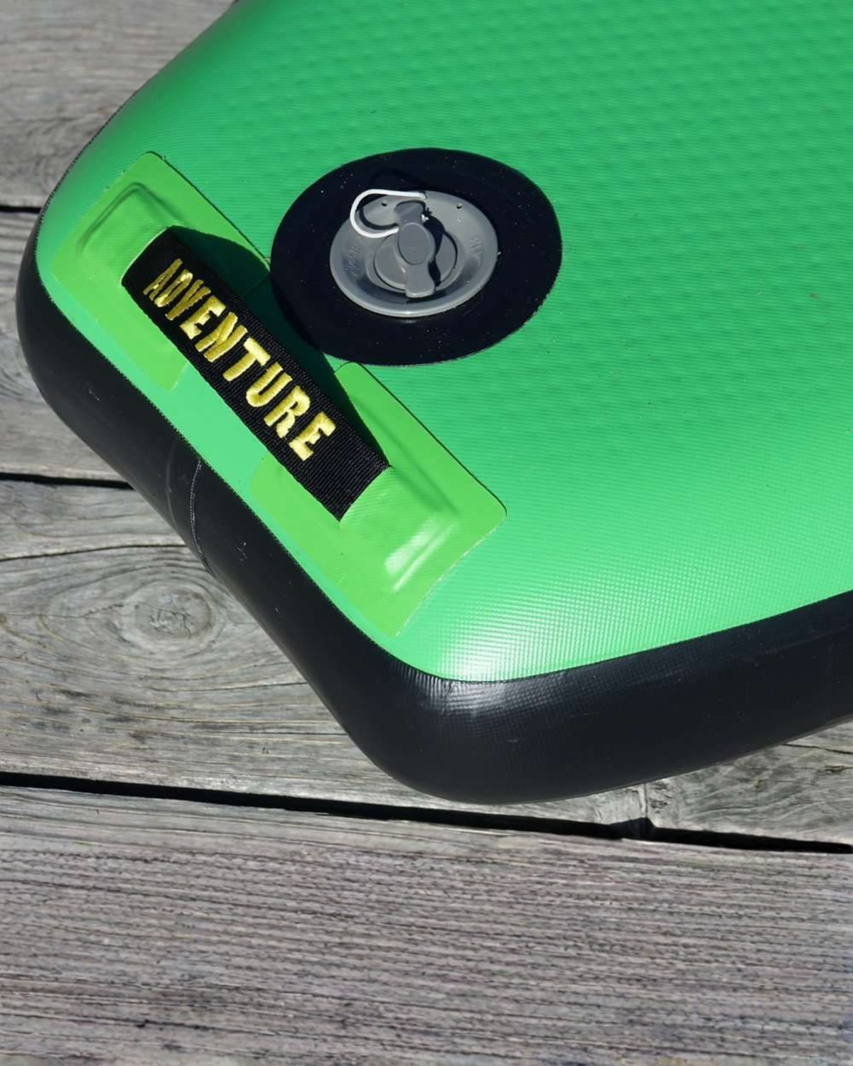 Close-up of a bright green inflatable board or bag with black trim, a circular black valve, and a black label reading 'ADVENTURE' in yellow text, placed on a gray wooden surface.