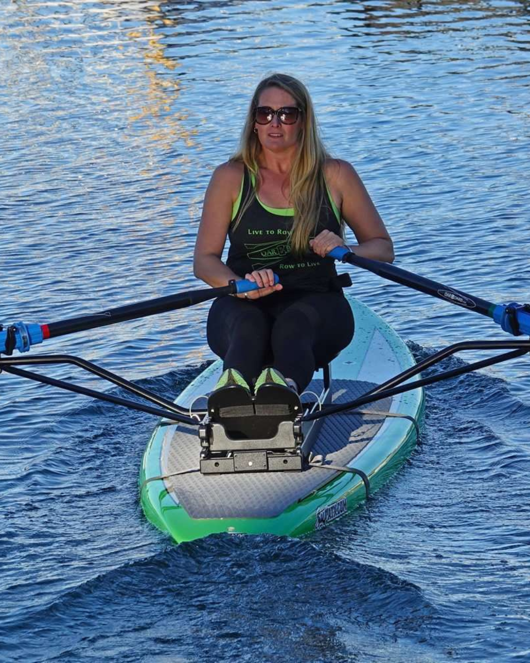 A woman rowing the oar board rowing kit attached to a green coloured hard paddleboard on calm, blue water.