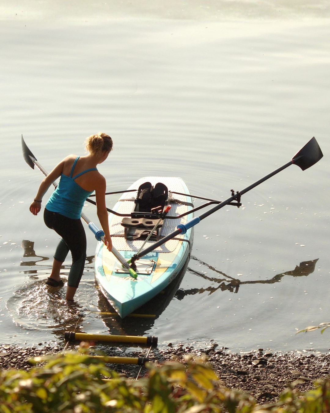 A woman in a blue tank top and black leggings stands in shallow water beside a light blue hard paddle board with the Oar Board rowing kit mounted