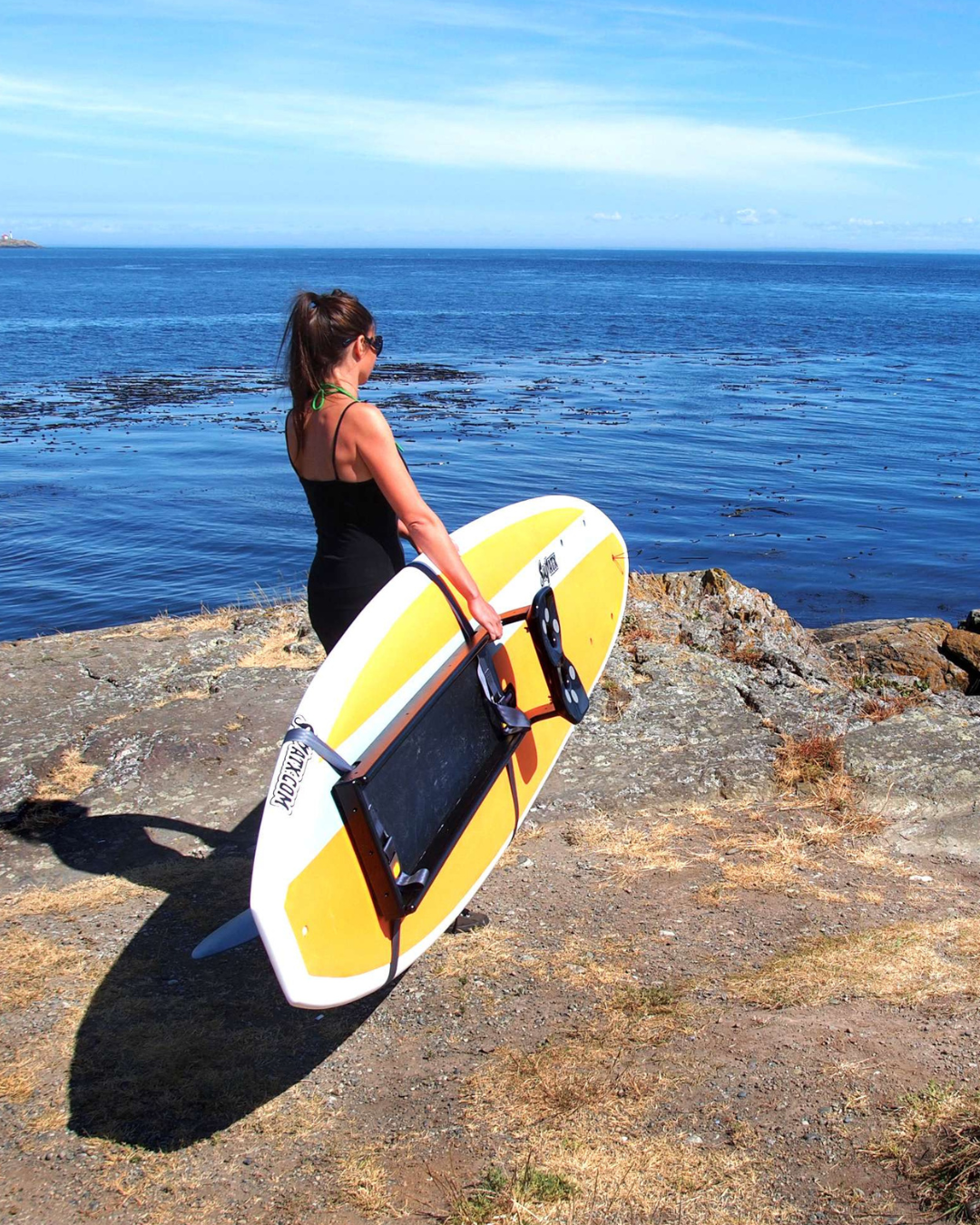 A woman in a black swimsuit stands on a rocky shoreline holding paddleboard with the rowing kit attached.