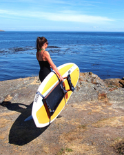 A woman in a black swimsuit stands on a rocky shoreline holding paddleboard with the rowing kit attached.